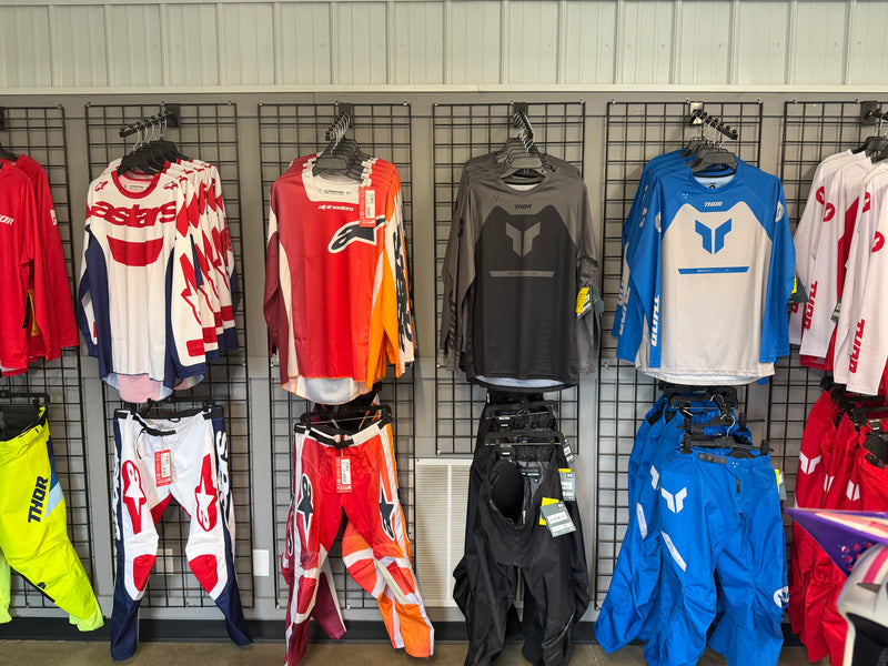 A retail display of motocross apparel hanging on black wire racks against a gray paneled wall. Visible brands include Alpinestars, Fly Racing, and Thor.
