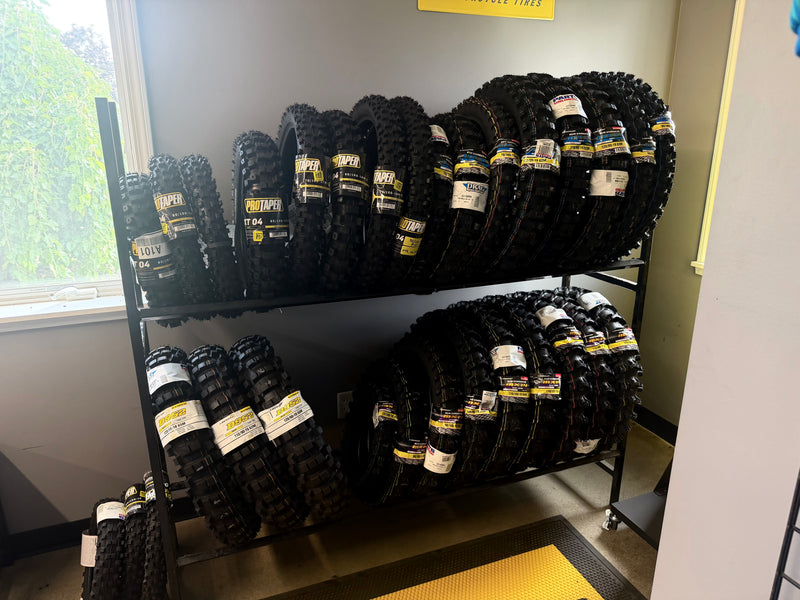 A set of Dunlop & Protaper off-road tires with white and yellow brand labels, displayed on a black metal rolling rack inside a store.