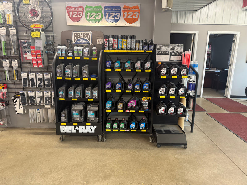 A Bel-Ray & Maxima branded display rack filled with various motor oils and power sports fluids, set inside a retail store with other products and store fixtures visible in the background.